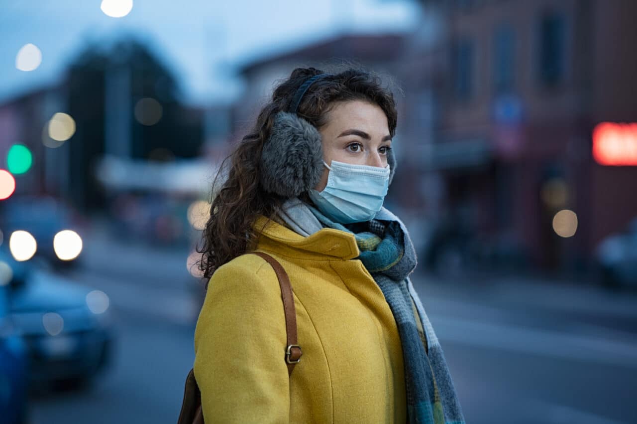Woman wearing a yellow coat, earmuffs and a face mask on a cold winter night.