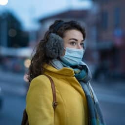 Woman wearing a yellow coat, earmuffs and a face mask on a cold winter night.