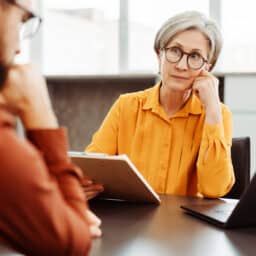 Older woman in yellow with hand on face, sitting across from a coworker who is talking to her